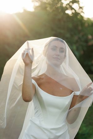 Bride in white gown and veil posing outdoors in golden sunlight with greenery background.
