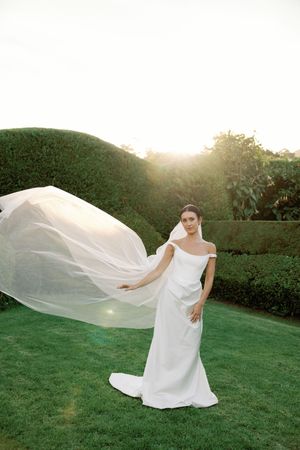 Bride in white off-shoulder gown with flowing veil caught in breeze, posing in lush garden at golden hour sunset.