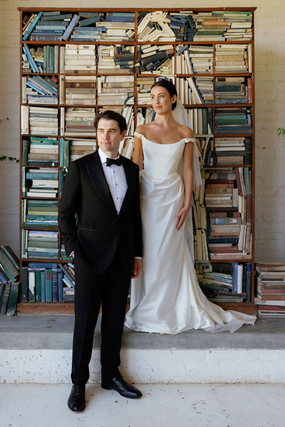 Bride in white off-shoulder gown and groom in black tuxedo pose before a rustic bookshelf backdrop.
