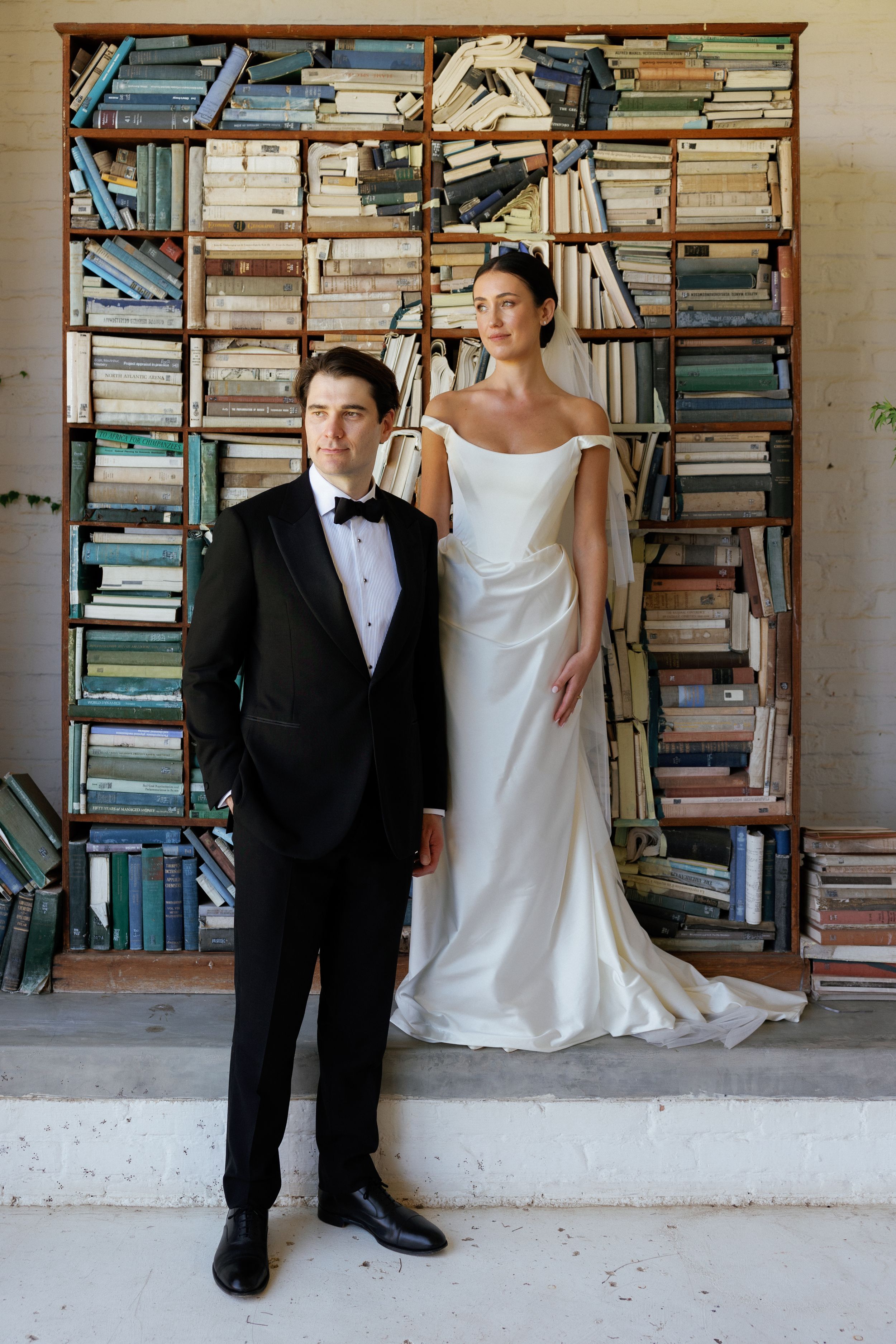 Bride in white off-shoulder gown and groom in black tuxedo pose before a rustic bookshelf backdrop.