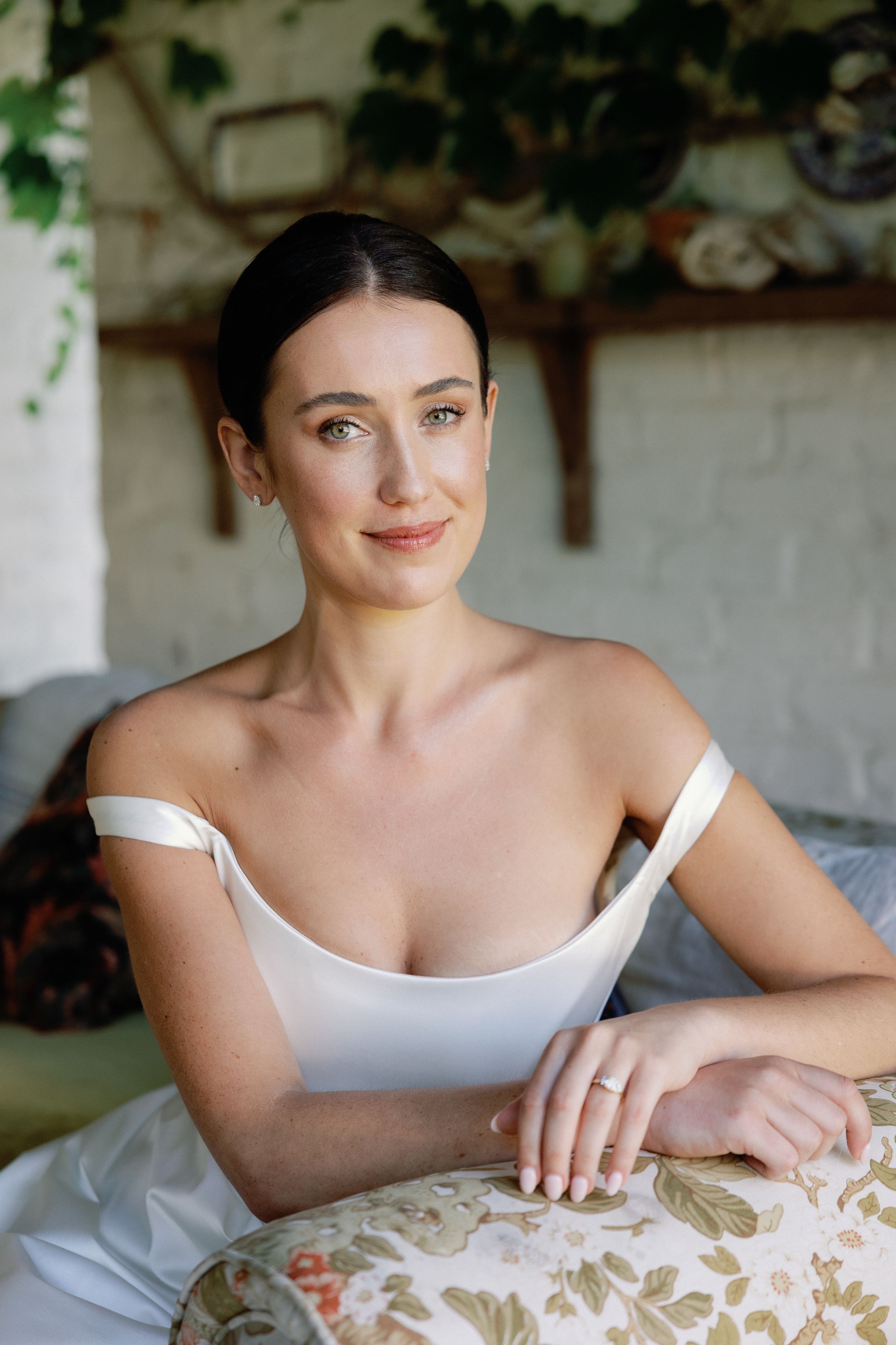 Elegant bride in white off-shoulder gown sitting gracefully, smiling softly with dark hair up in a chic updo.