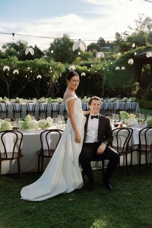 Bride in white gown and groom in tuxedo pose beside an elegant outdoor wedding reception table with floral arrangements.