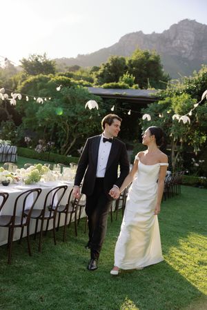 Bride and groom walk hand-in-hand on lush lawn beside elegant outdoor reception tables with mountain backdrop.