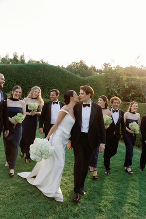 Bride and groom kiss while walking with wedding party in navy tuxedos and dresses on lush green lawn at sunset.
