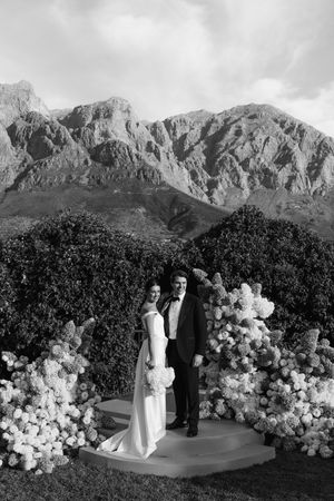 Black and white wedding photo of bride and groom standing before floral arch with dramatic mountain backdrop.