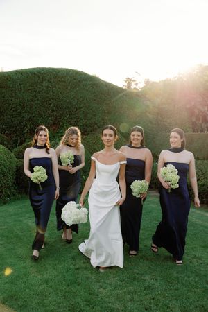 Bride in white gown walks with four bridesmaids in navy dresses holding green bouquets on lush garden lawn at sunset.