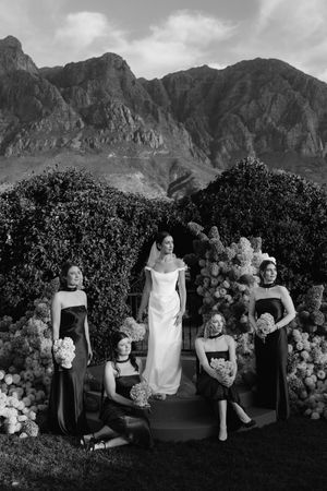 Black and white photo of a bride in white gown with four bridesmaids in dark dresses, mountains and florals backdrop.