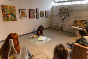 Artist demonstrates painting techniques on gallery floor surrounded by observers, with colorful artwork displayed on white walls.