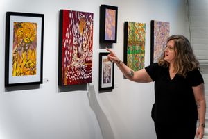 Woman in black outfit pointing at colorful abstract paintings displayed on a white gallery wall.