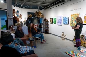 Artist giving a gallery talk to seated audience in a colorful art gallery with paintings on white walls.