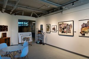 Modern art gallery interior with white walls displaying framed artwork, Eames chairs, and industrial ceiling.