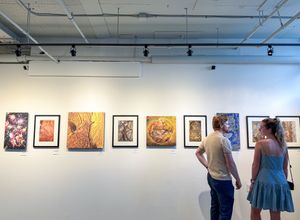 Two visitors viewing colorful framed artwork displayed on white gallery walls under track lighting.