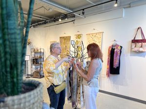 Two people browsing colorful scarves and jewelry in a bright boutique art gallery shop with wall hangings and accessories.
