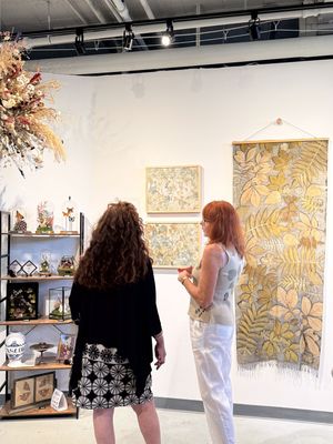 Two women viewing botanical artwork and floral wall hangings in a bright art gallery with wooden shelving displays.