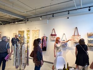 Visitors browse handmade bags and textile art displayed on white gallery walls during an art show opening event.