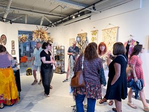 People browsing colorful artwork and textiles at an indoor art gallery opening event.