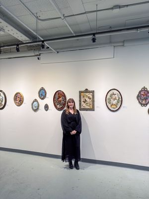 Woman in black dress stands before gallery wall displaying ornate framed botanical and nature artwork in oval and rectangular frames.