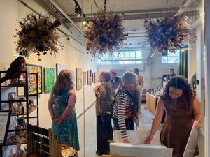 Visitors browse artwork and floral installations at an art gallery opening event with colorful paintings on white walls.