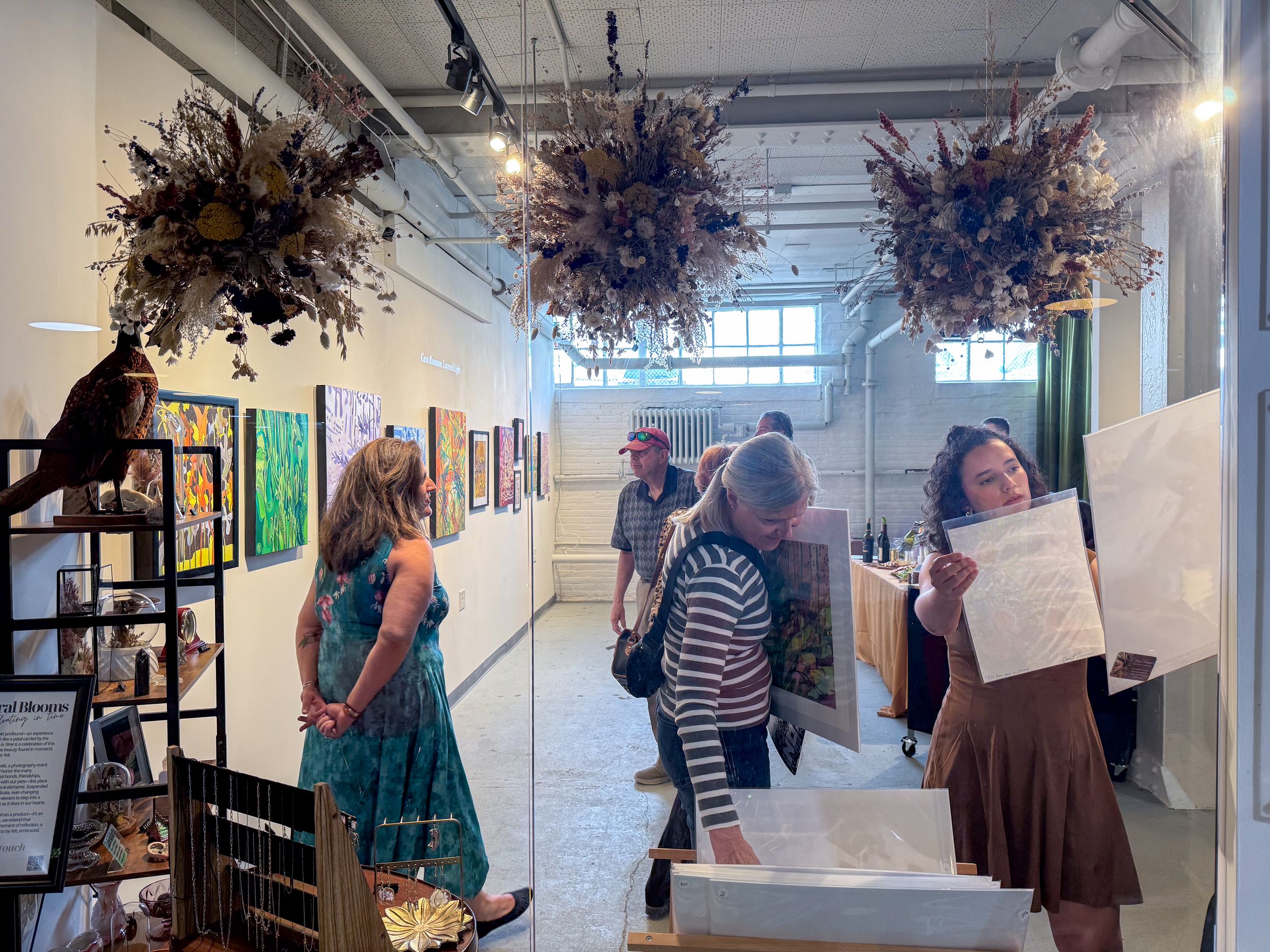Visitors browse colorful artwork in a gallery with dried floral installations hanging from the ceiling.