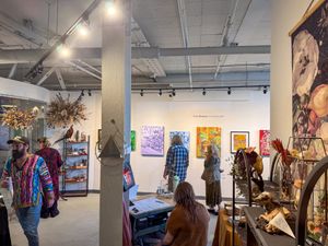Visitors browse colorful paintings and sculptures in a modern art gallery with exposed ceilings and track lighting.