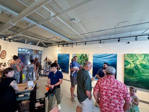 Visitors browse vibrant ocean-themed paintings at a contemporary art gallery opening event with track lighting.