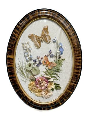 Oval framed botanical art with pressed flowers, ferns, and two butterflies on textured white background in tortoiseshell frame.