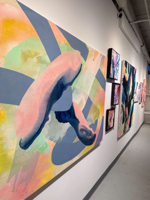 Colorful abstract paintings displayed on a white gallery wall, featuring large pink and blue brushstrokes and smaller framed artworks.