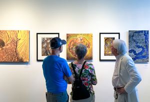 Three visitors admire colorful framed artwork displayed on a white gallery wall during an art exhibition.