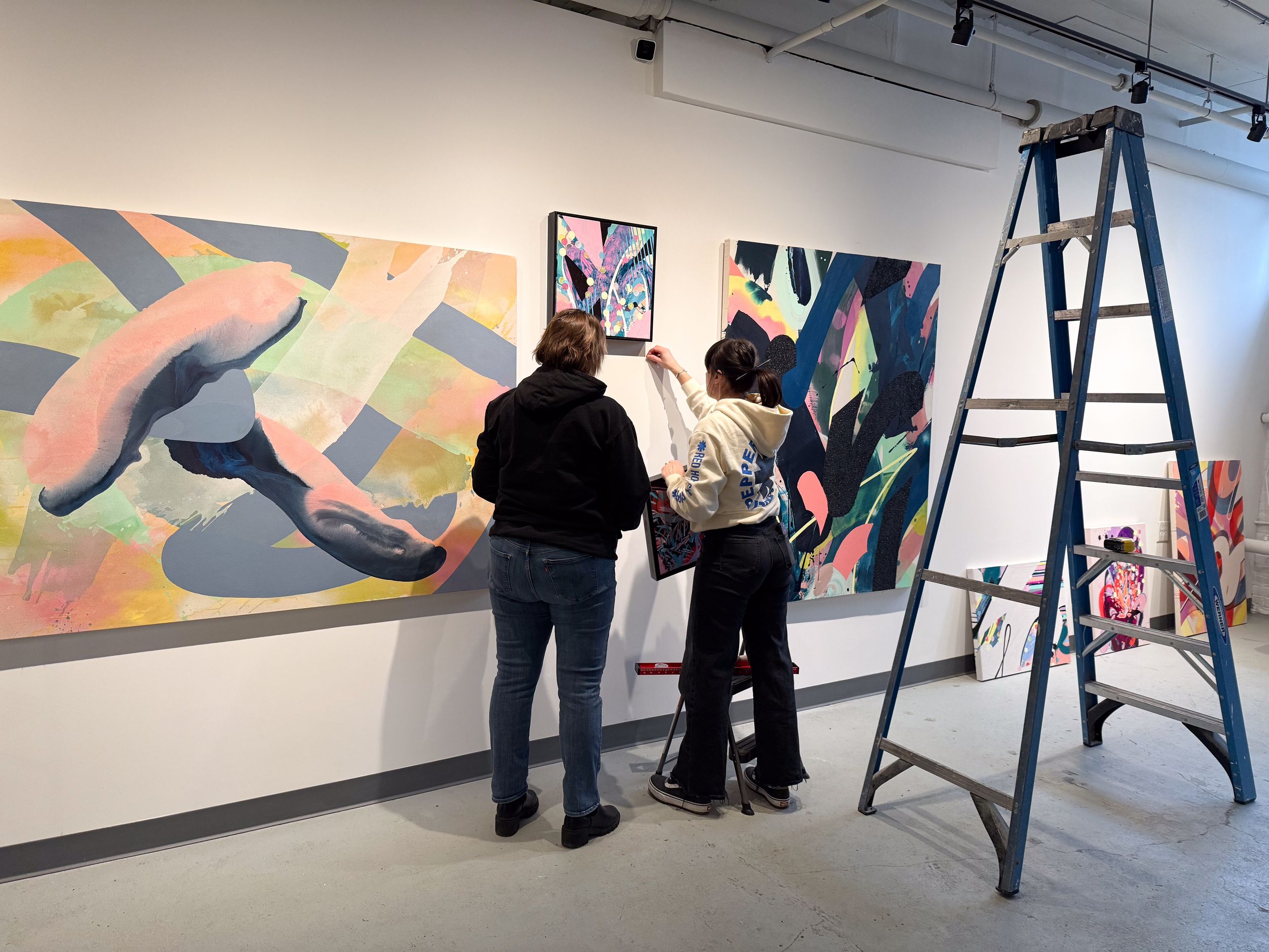 Two people hang colorful abstract paintings on a white gallery wall next to a ladder during an art exhibition setup.