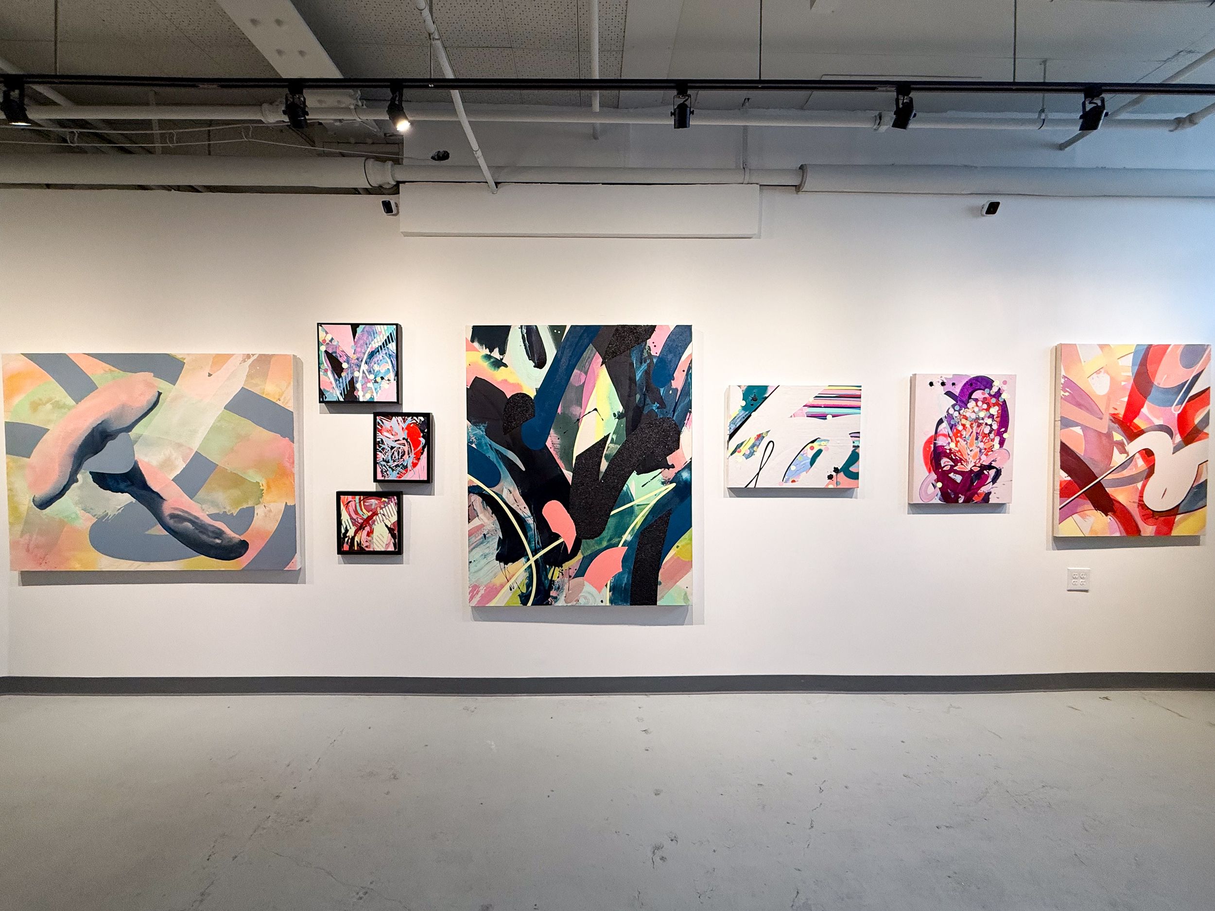 Colorful abstract paintings displayed on a white gallery wall in a modern art exhibition space with concrete floors.