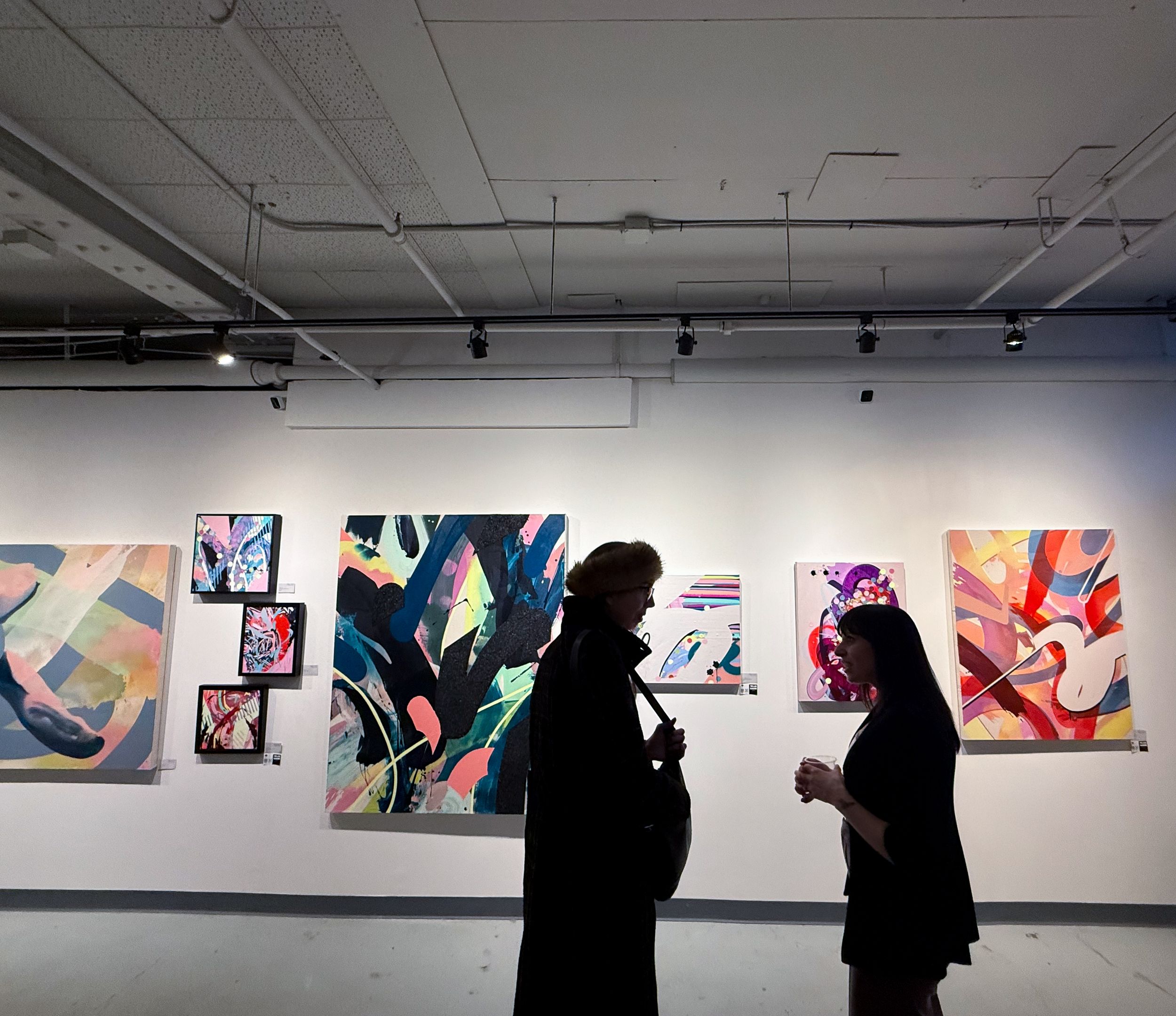 Two silhouetted visitors viewing colorful abstract paintings displayed on white gallery walls at an art exhibition.