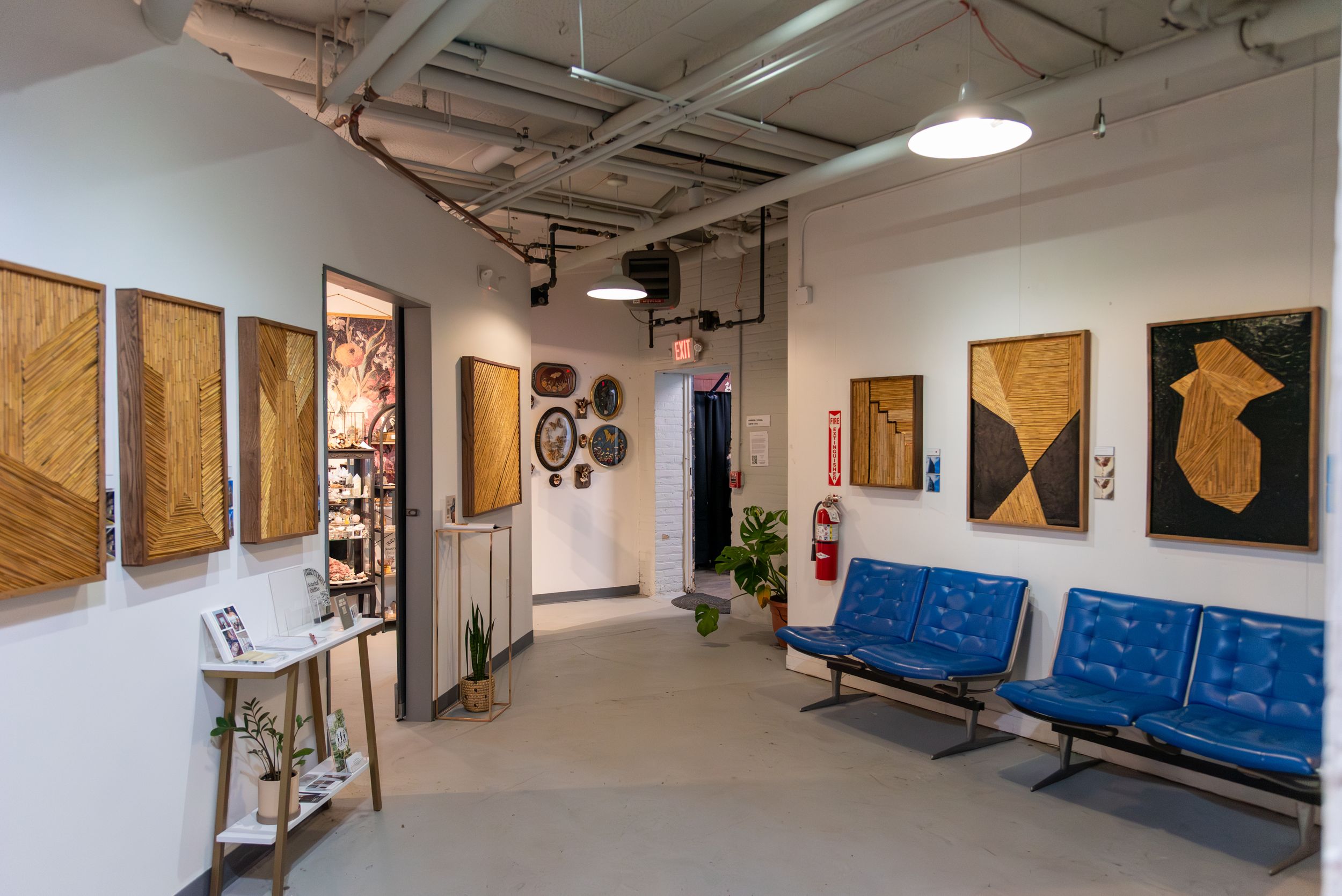 Modern art gallery lobby with wooden artwork panels, blue chairs, white walls, exposed pipes, and decorative plants.