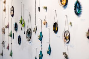 Colorful handmade jewelry and circular art pieces displayed on a white gallery wall, featuring teal geometric pendants.