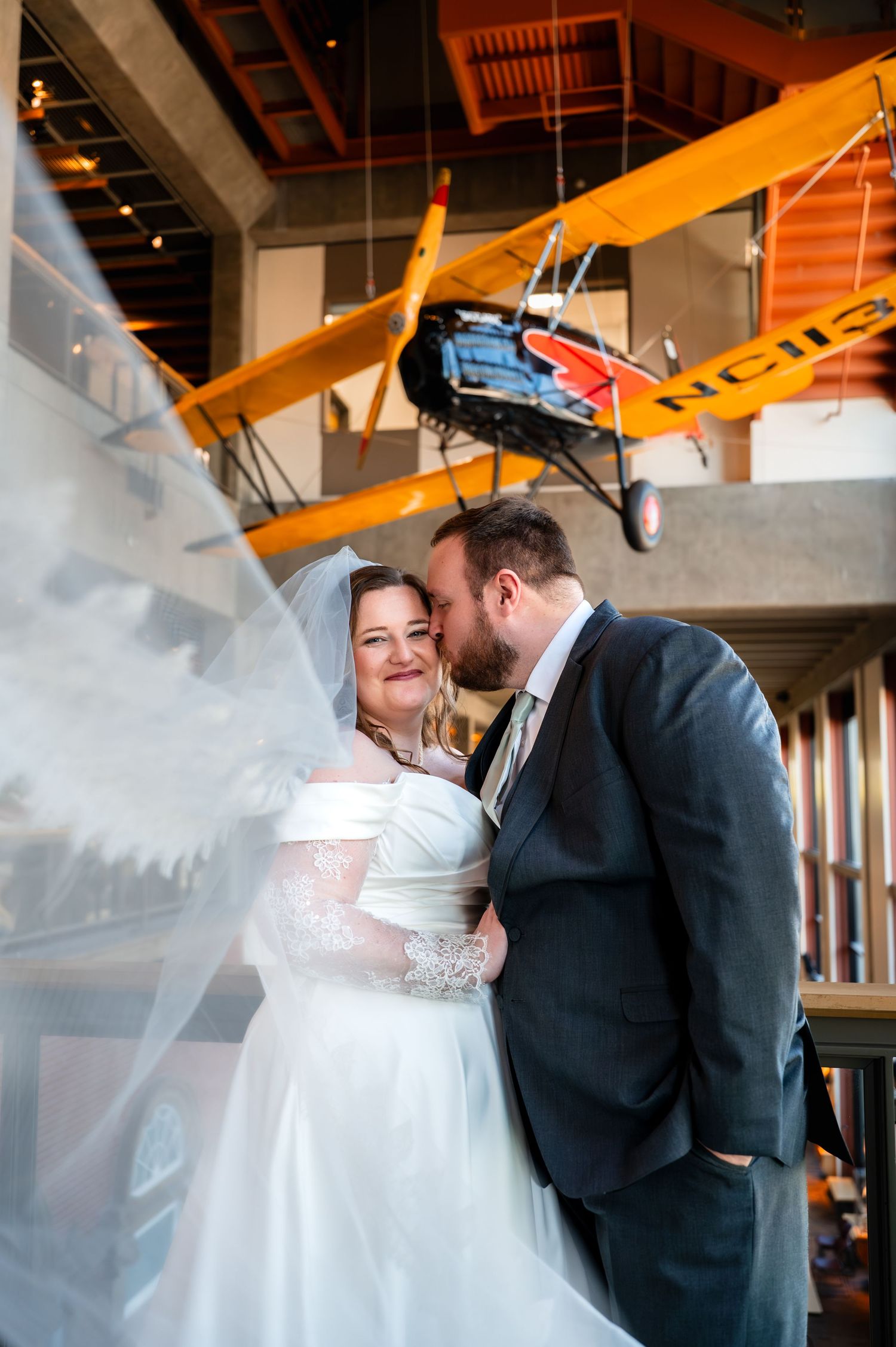 Bride and groom share a romantic moment in a museum with a vintage yellow biplane hanging overhead, veil flowing beautifully.