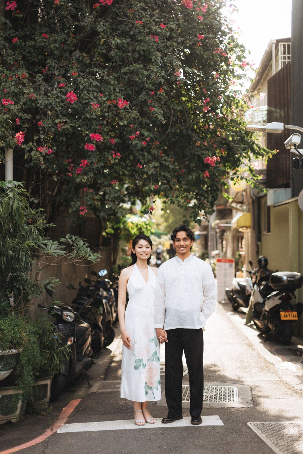 Couple in white attire holding hands on a charming alley street under blooming pink bougainvillea trees.