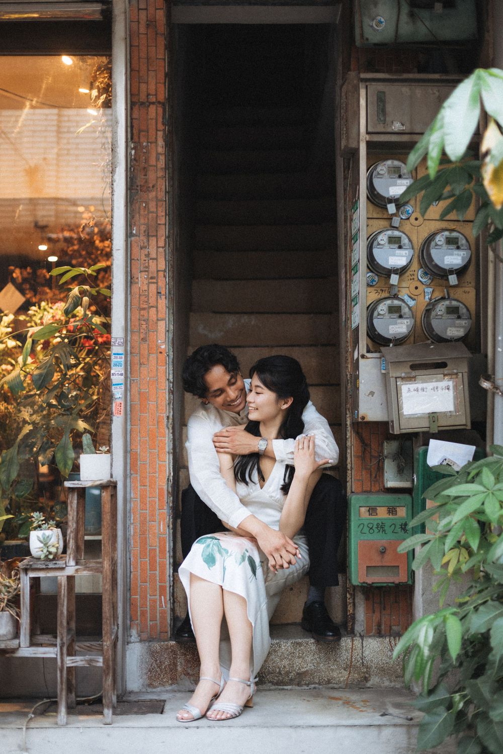 Couple embracing romantically in a vintage doorway with electric meters, surrounded by lush plants in an Asian neighborhood.
