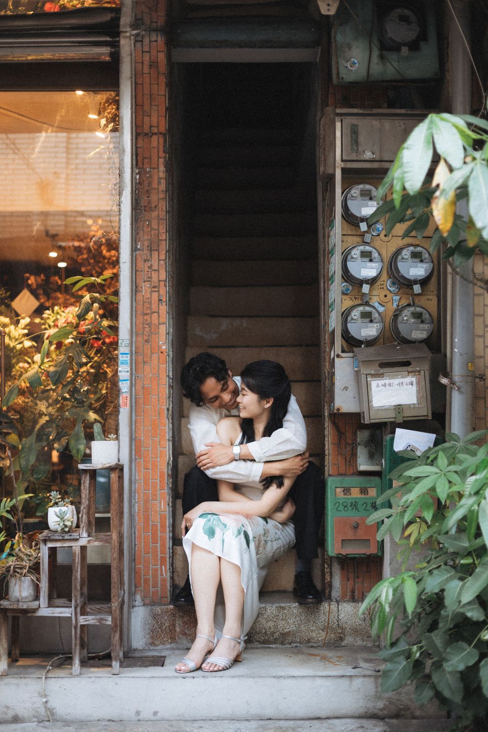 Couple embracing romantically on vintage building steps surrounded by lush plants and old utility meters in Asia.