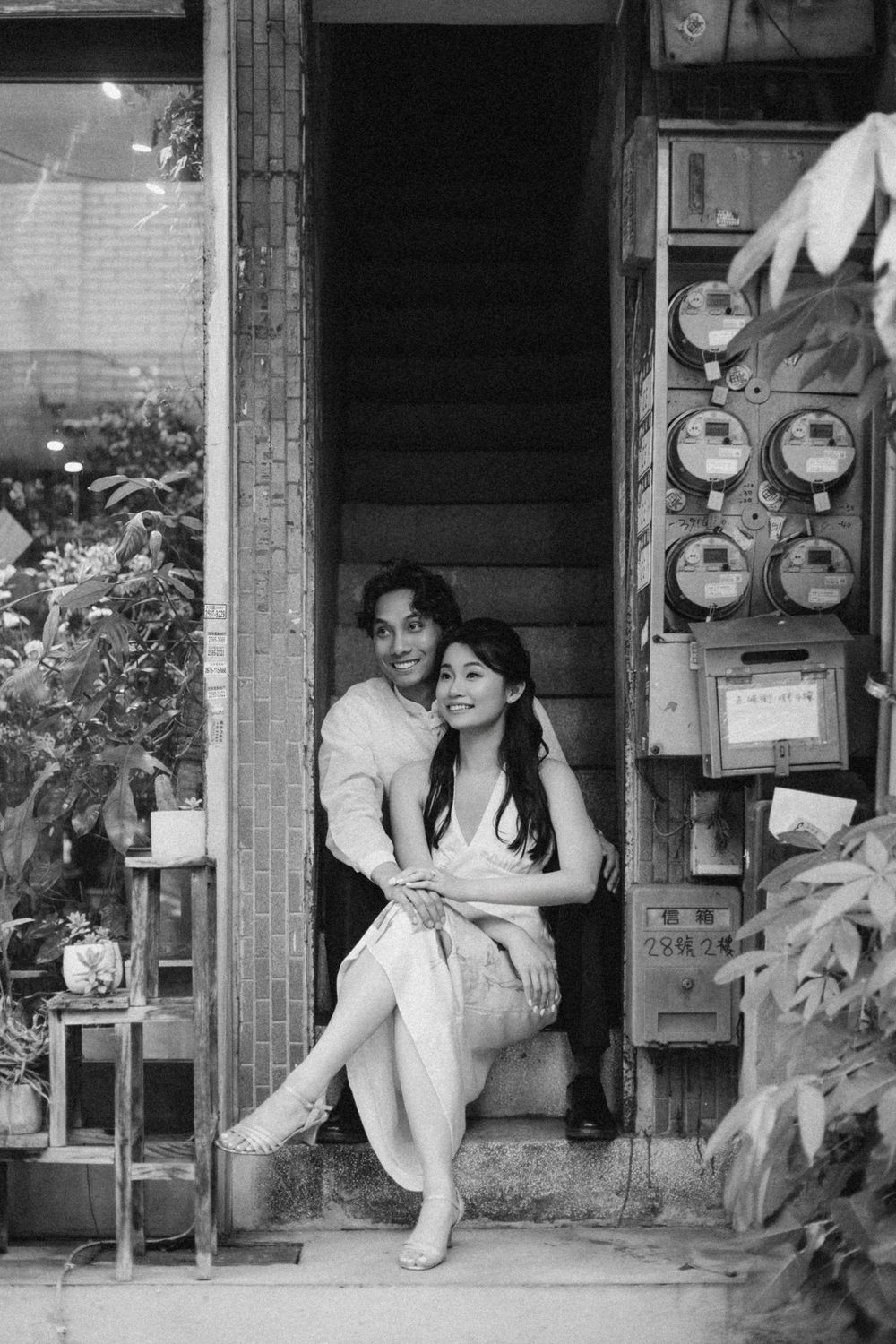 Black and white photo of a couple sitting in a doorway, embracing warmly outside a vintage shop.