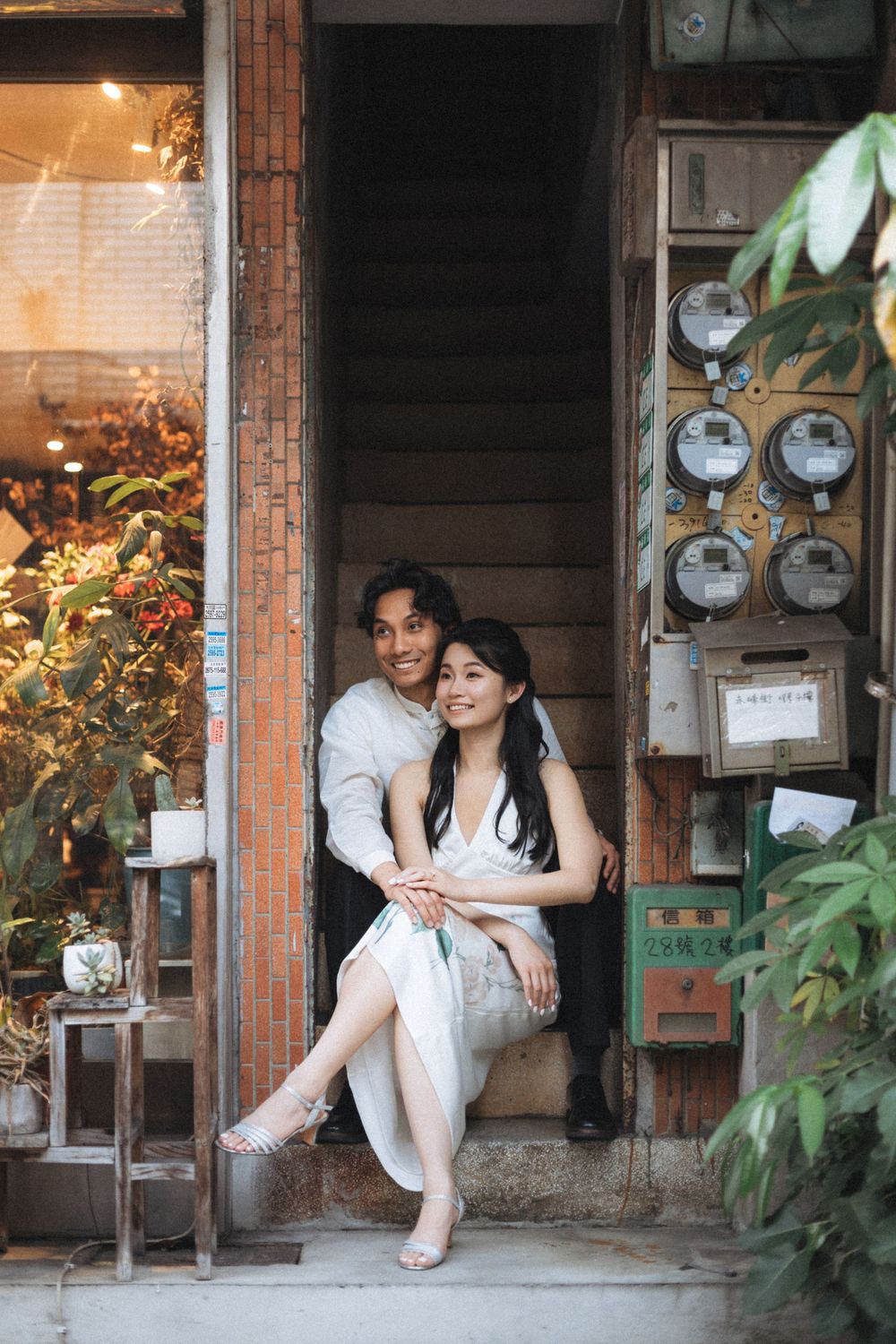 Couple sitting together on vintage shop steps, woman in white dress, man in gray shirt, surrounded by plants and utility meters.