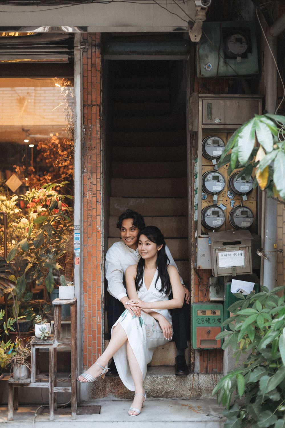 A couple sits together on vintage shop steps surrounded by lush plants and old utility meters.
