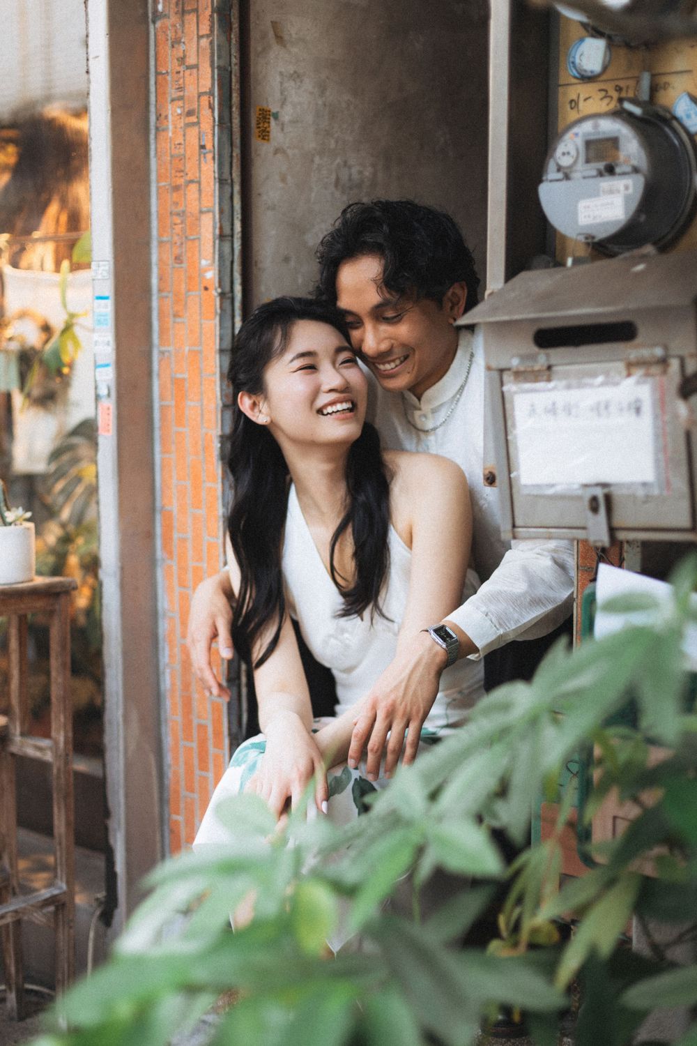 Happy Asian couple embracing and laughing together outside a vintage building with lush green plants.