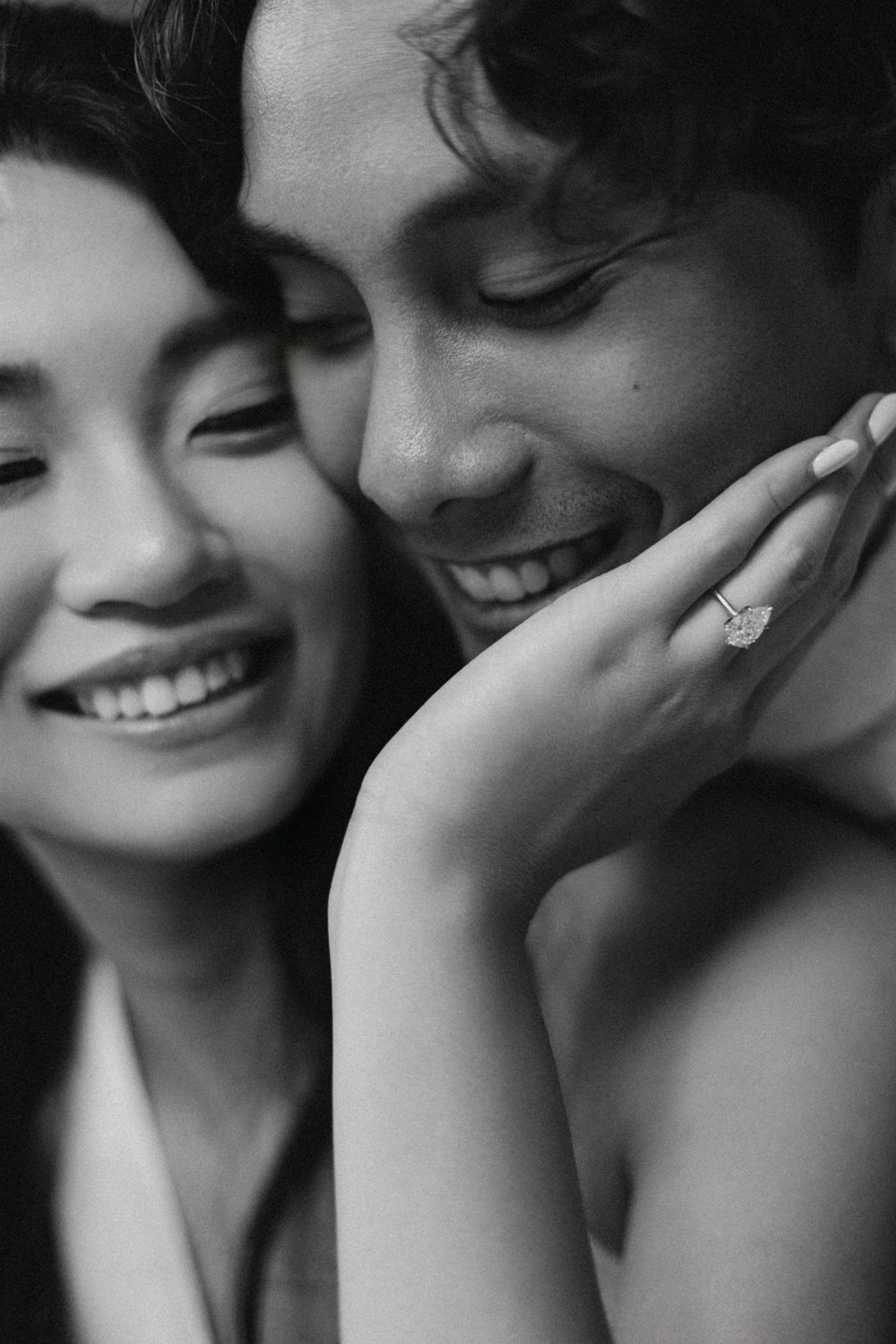 Black and white photo of a smiling couple, woman displaying a diamond engagement ring while embracing tenderly.