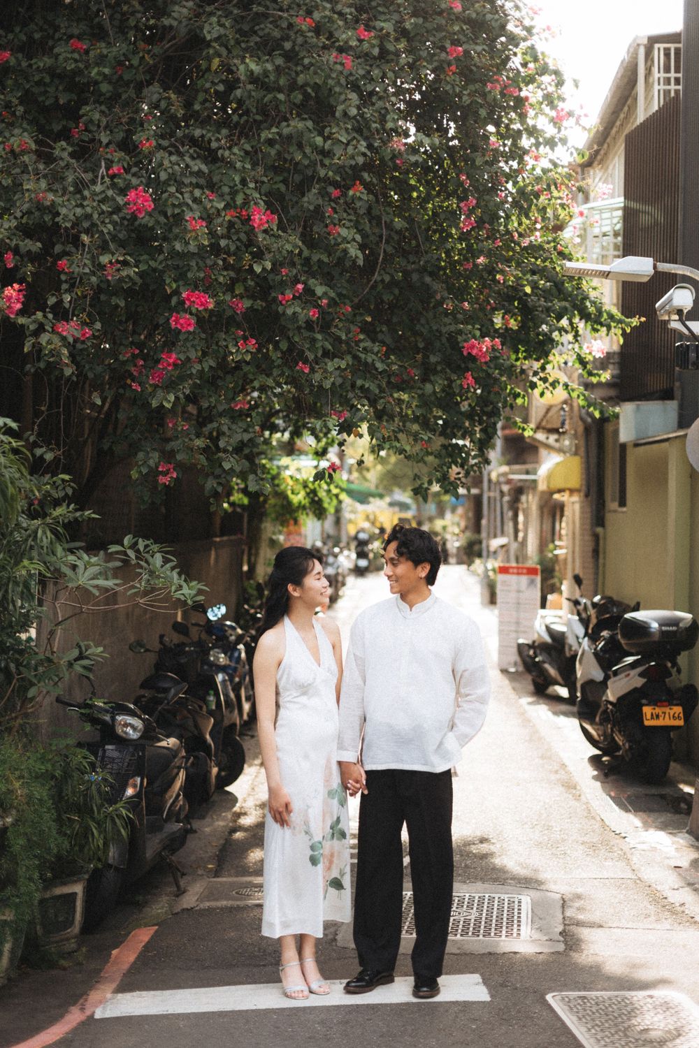 Couple in white attire holding hands on a narrow alley lined with motorcycles and blooming bougainvillea trees.