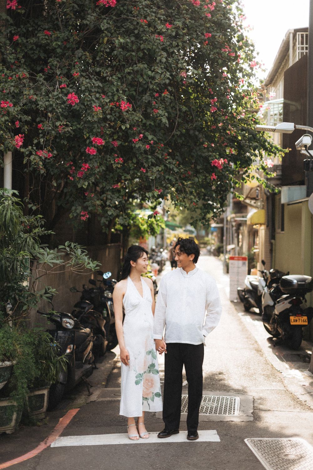 Couple standing on a crosswalk in a charming alley with blooming pink flowers and parked motorcycles.