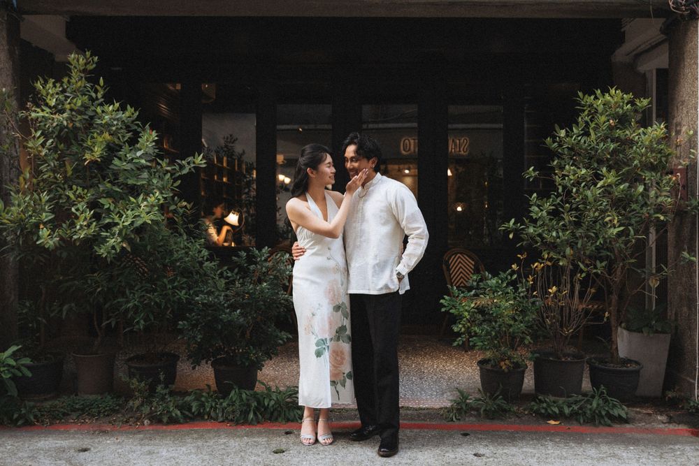 Couple in elegant attire embracing outside a rustic building with lush potted plants.