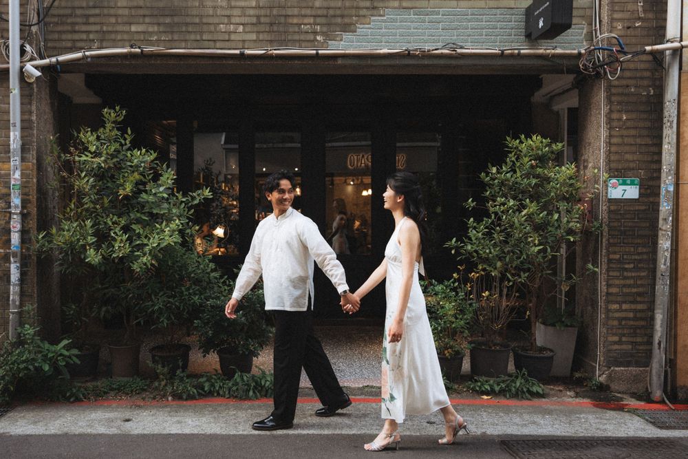 Couple in elegant attire holding hands outside a vintage shop, man in white shirt, woman in white dress.