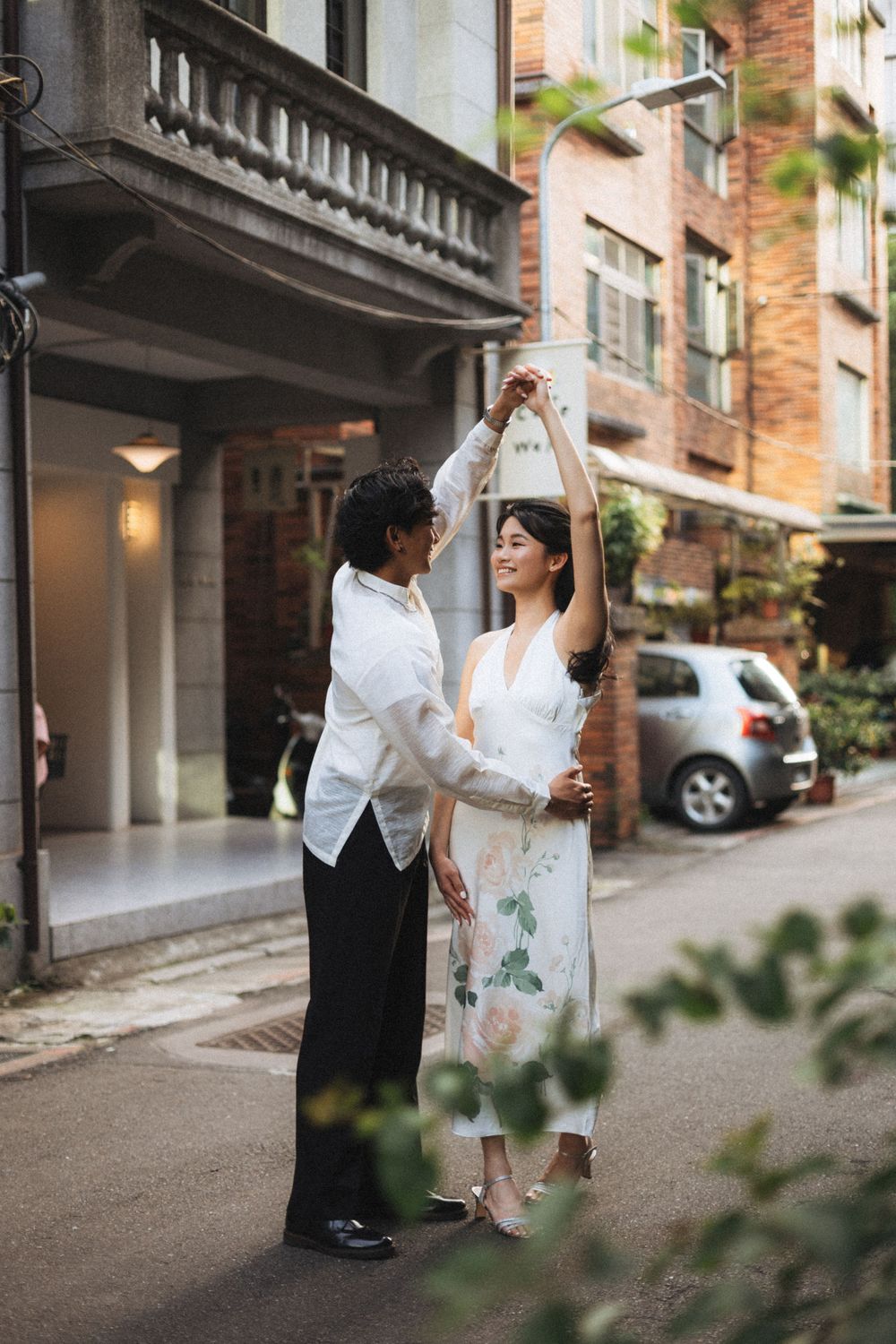 Couple dancing together on a charming urban street, woman in floral dress being twirled by man in white shirt.