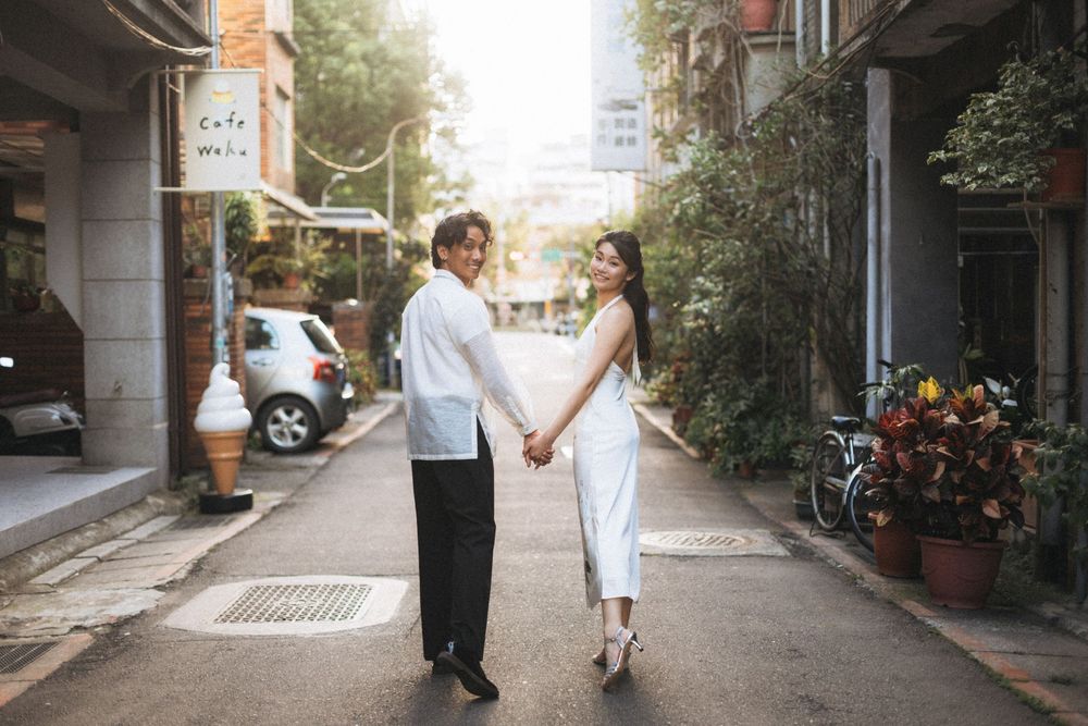 Couple holding hands while posing on a charming urban alley, dressed in elegant white and black outfits.