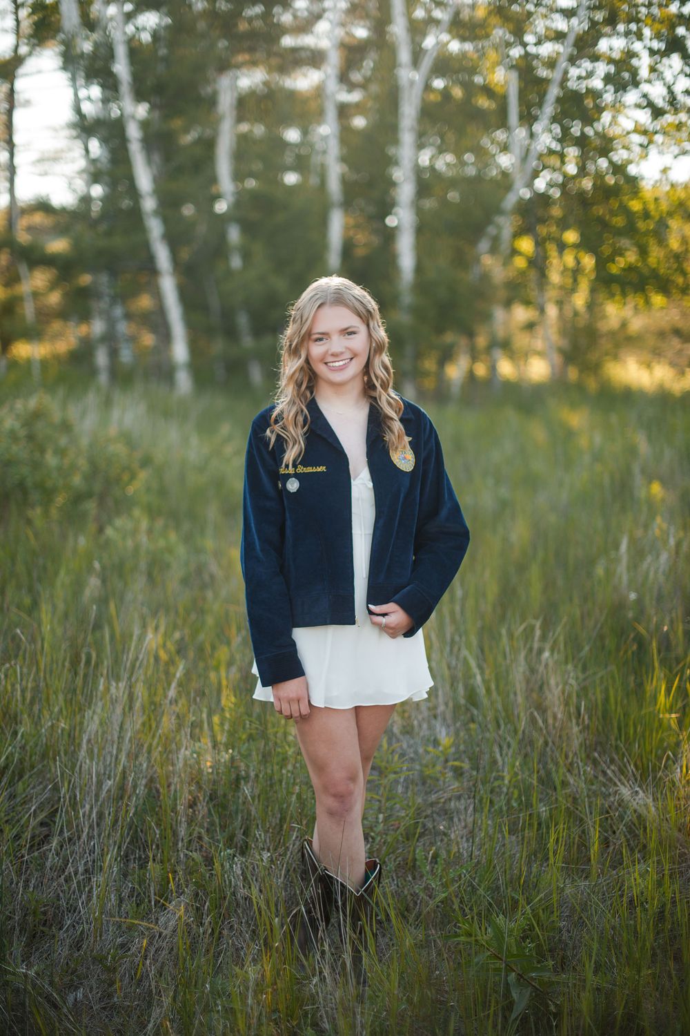 Smiling blonde teen in navy jacket and white dress poses in sunlit meadow with birch trees.
