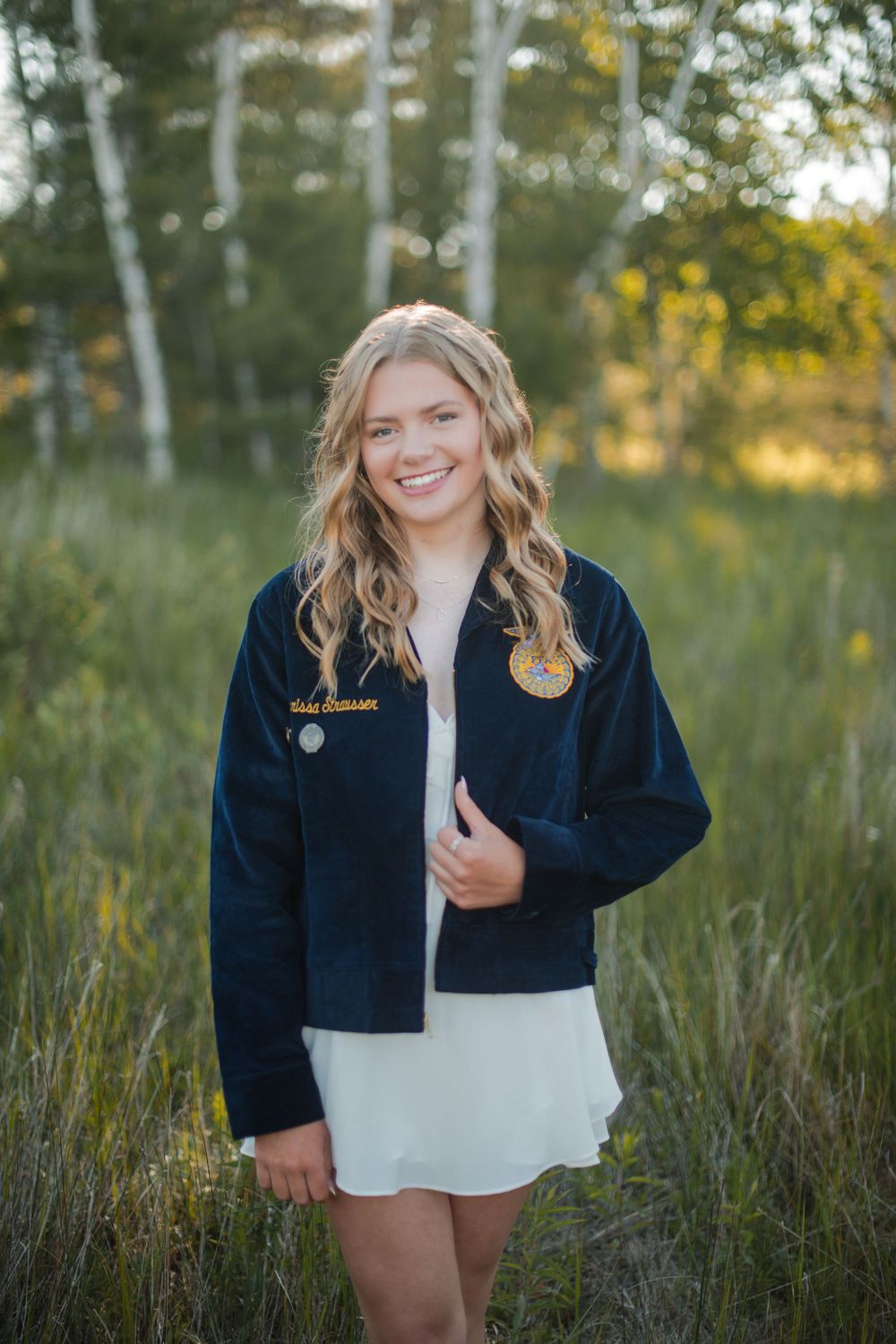 Smiling teenage girl in FFA jacket and white dress standing in a sunlit meadow with birch trees.
